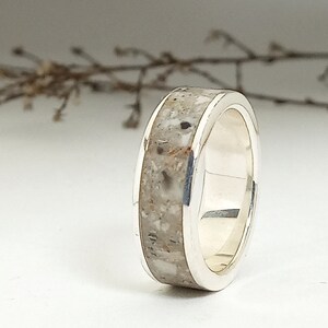 Puede incluir: Un anillo de banda de plata con una incrustación texturizada de color gris claro. El anillo está hecho de metal y tiene un acabado pulido.