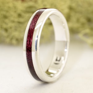 Könnte beinhalten: Ein silberner Ring mit einer dünnen Einlage aus dunkelrotem Holz. Der Ring ist glatt und poliert.