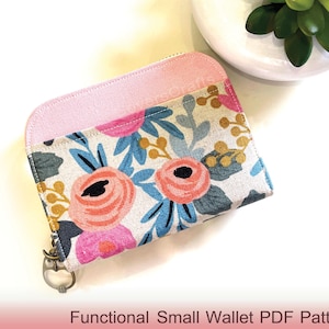 Puede incluir: Una pequeña cartera con estampado floral con la parte superior rosa y cierre de cremallera. La cartera presenta un diseño de rosas rosas, hojas azules y detalles dorados sobre un fondo crema. El texto "Functional Small Wallet PDF Pattern" está en la parte inferior.