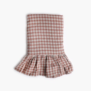 May include: A folded bed skirt in a terracotta and white gingham pattern. The fabric has a slightly wrinkled texture. The bed skirt features a ruffled edge at the bottom.