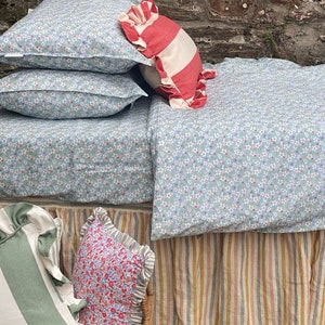 May include: A bed with a blue floral patterned duvet cover and a striped bed skirt. There are several pillows on the bed, including a red and white striped pillow with ruffled edges. The bed is against a stone wall with green vines and white flowers.