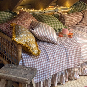 May include: A brown and white gingham daybed with a ruffled skirt and a variety of patterned pillows. The daybed is decorated with fairy lights and a small wooden stool.
