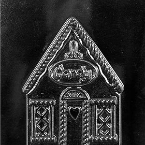 Puede incluir: Un molde de metal negro para hacer caramelos con forma de casa de jengibre. El molde tiene un diseño de cuerda alrededor de los bordes y las ventanas, y las palabras "Candy" están en relieve en la parte delantera.