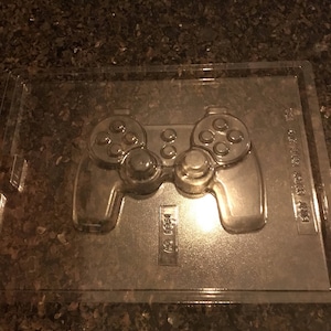 May include: A clear plastic mold for making chocolate video game controllers. The mold has a detailed design of a controller with buttons and a directional pad.