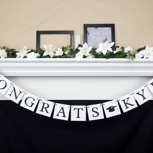 May include: A white banner with black letters spelling "Congrats Kyle" with a graduation cap in between "grats" and "Kyle". The banner is strung with green ribbon and is hanging above a fireplace.