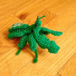 May include: A green crocheted praying mantis with pipe cleaner legs. The mantis is sitting on a wooden floor.