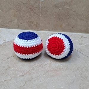 May include: Two spherical crochet balls featuring a striped pattern of red, white, and blue yarn. The left ball shows horizontal bands of color, while the right ball displays a circular red center surrounded by concentric rings of white and blue yarn.