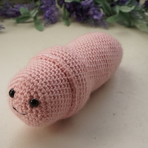 May include: A pink crocheted worm with black eyes. The worm is curled up on a white surface.