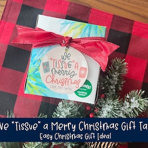 May include: A gift box with a red ribbon and a round tag that reads "We 'Tissue' a Merry Christmas." The box is on a red and black plaid surface, with a sprig of evergreen and a box of tissues visible.