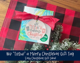 May include: A gift box with a red ribbon and a round tag that reads "We 'Tissue' a Merry Christmas." The box is on a red and black plaid surface, with a sprig of evergreen and a box of tissues visible.