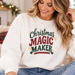May include: A white crewneck sweatshirt featuring the text Christmas MAGIC MAKER in a retro-style font. The word Christmas is in dark green, MAGIC is in red, and MAKER is in dark green, accented by small yellow star icons throughout the design.