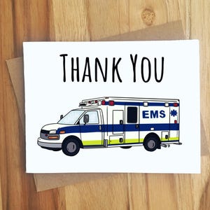May include: A white greeting card with the words "THANK YOU" in black lettering. The card features an illustration of a white, blue, and yellow ambulance with the letters "EMS" on the side. The card is on a brown envelope.