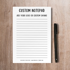 May include: A white custom notepad with horizontal black lines and the text "CUSTOM NOTEPAD ADD YOUR LOGO OR CUSTOM SAYING". A black pen rests on a wooden surface next to the notepad. The bottom of the notepad has the text "BAD JOKES BY GOOD PEOPLE - WWW.SKPINK.COM - @SKP_INK".