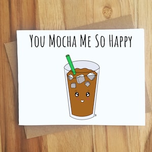 May include: A white card with the text "You mocha me so happy" and a cartoon illustration of a glass of iced coffee with a green straw and ice cubes. The coffee has a smiling face.