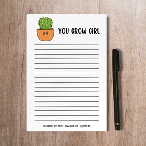 May include: A lined notepad with the phrase "YOU GROW GIRL" and a cartoon cactus in a pot. A black pen rests on the right side of the notepad. The notepad has the text "BAD JOKES BY GOOD PEOPLE - WWW.SKPINK.COM - @SKP_INK" at the bottom.