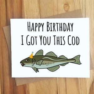May include: A white greeting card with the black text Happy Birthday I Got You This Cod. Below the text is a cartoon illustration of a green and grey cod fish wearing a small yellow party hat with orange polka dots and a blue pom-pom.