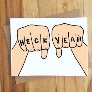 May include: A white greeting card with a black line drawing of two fists with the words "HECK YEAH" spelled out on the knuckles.