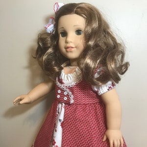May include: A doll wearing a red and white dress with a white lace trim. The dress has a floral pattern on the bottom half. The doll has long brown hair and green eyes.