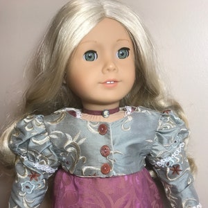 May include: A doll wearing a light blue and silver embroidered jacket with brown buttons and a pink floral skirt.