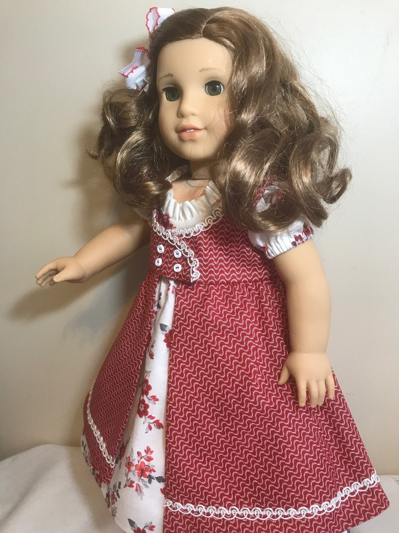 May include: A doll wearing a red and white dress with a white lace trim. The dress has a floral pattern on the bottom half. The doll has long brown hair and green eyes.