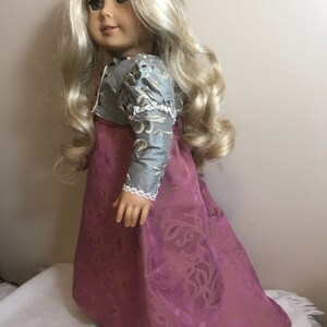 May include: A doll wearing a long, pink dress with a floral pattern and a blue and silver jacket. The doll has long blonde hair and blue eyes.