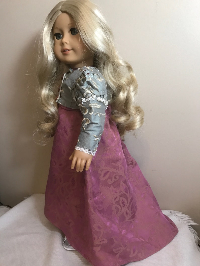May include: A doll wearing a long, pink dress with a floral pattern and a blue and silver jacket. The doll has long blonde hair and blue eyes.