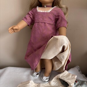May include: A doll wearing a pink and gold dress with a white lace collar and silver shoes. The doll is holding a white fabric skirt in her hands.