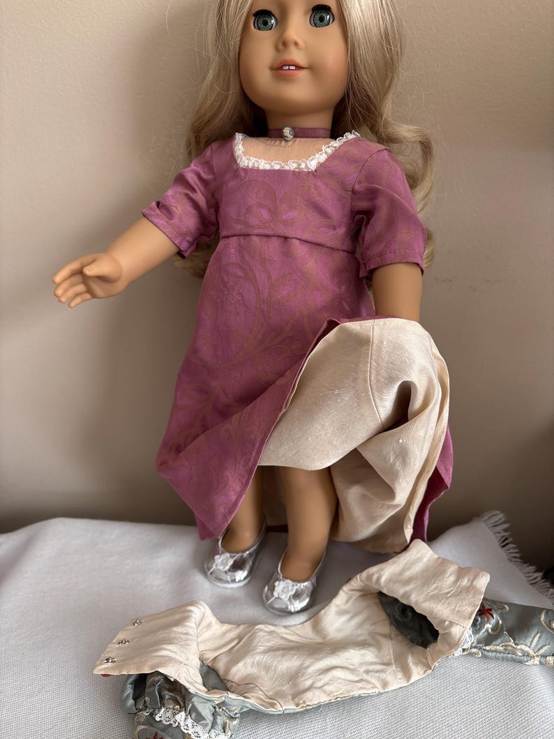 May include: A doll wearing a pink and gold dress with a white lace collar and silver shoes. The doll is holding a white fabric skirt in her hands.