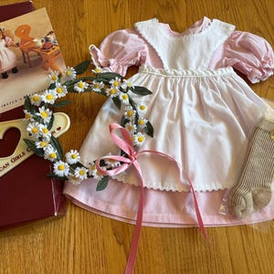 May include: A pink and white dress with a white apron and a pink ribbon. The dress has a gingham pattern on the bottom. A daisy flower crown and a pair of brown socks are also included. The text "The American Girls Collection" and "Kirsten's Birthday" are visible on the red box.
