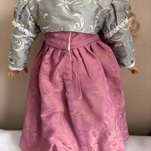 May include: A doll wearing a pink dress with a floral pattern and a light blue jacket with a floral pattern. The jacket has white lace trim on the sleeves.