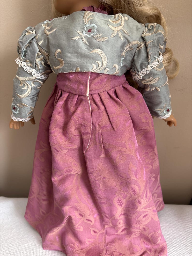 May include: A doll wearing a pink dress with a floral pattern and a light blue jacket with a floral pattern. The jacket has white lace trim on the sleeves.