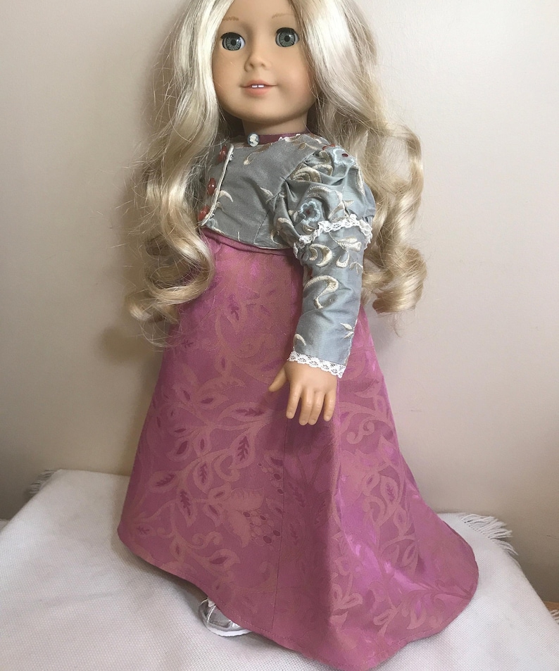 May include: A doll wearing a long, pink dress with a floral pattern. The dress has a gray bodice with a floral pattern and white lace trim. The doll has long blonde hair and blue eyes.