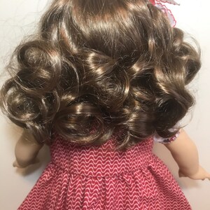 May include: A doll with long, brown, curly hair. The doll is wearing a red dress with a white pattern.