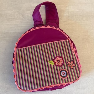 May include: A magenta backpack with a handle and a striped front pocket. The pocket features vertical stripes in shades of green, pink, and brown. The backpack is decorated with floral appliqués and a circular design. The edges are trimmed with a scalloped pink border.