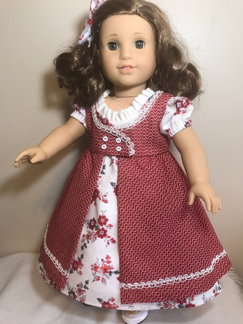 May include: A red and white dress with a floral print and white lace trim. The dress is worn by a doll with brown hair and green eyes.