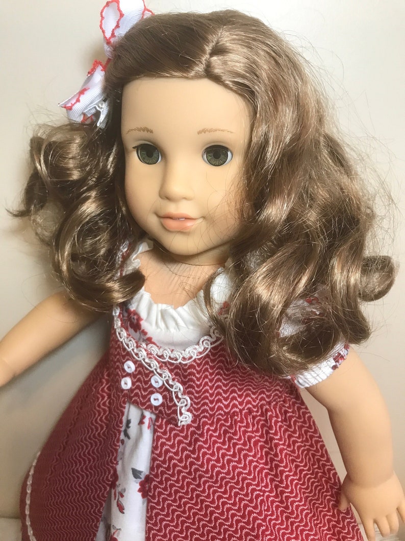 May include: A doll with long brown curly hair wearing a red and white dress with a white lace trim. The doll has green eyes and is wearing a white bow with red accents in her hair.