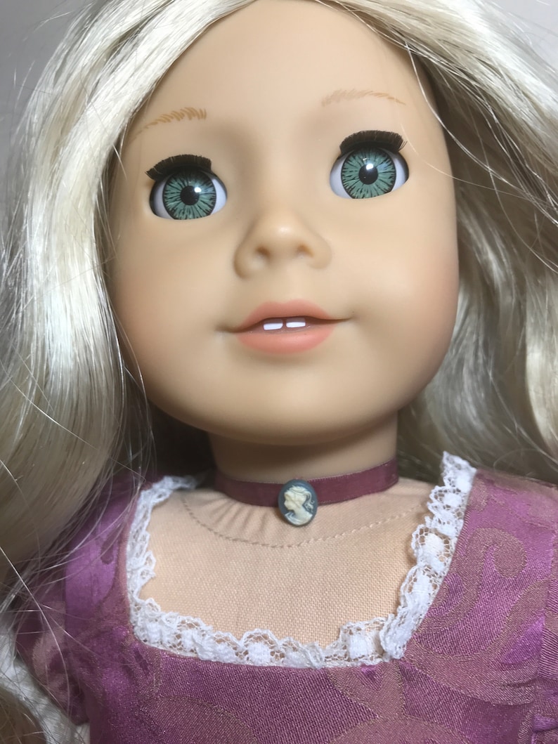 May include: A doll with blonde hair wearing a purple dress with white lace trim. The doll has green eyes and is wearing a dark red choker with a cameo pendant.