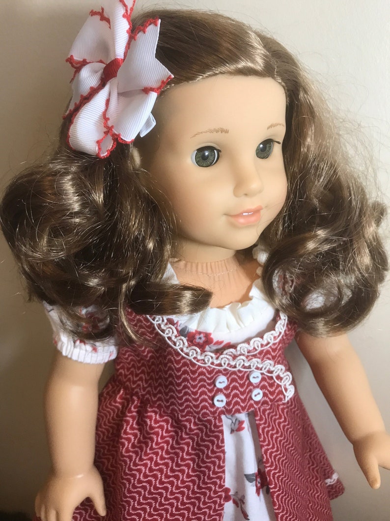 May include: A doll with long brown curly hair wearing a red and white dress with a white lace trim. The doll has green eyes and is wearing a white bow with red trim in her hair.