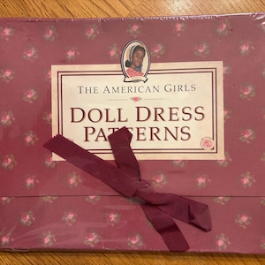 May include: A boxed set of "The American Girls Doll Dress Patterns" is wrapped in clear plastic. The box is a deep rose color with a floral pattern and a burgundy ribbon. The box features a portrait of a girl.