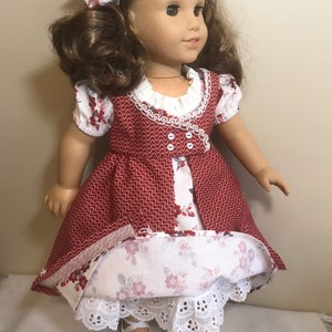 May include: A doll wearing a red and white dress with a floral print. The dress has a white lace trim and a white ruffle collar. The doll is wearing white shoes with gold accents.