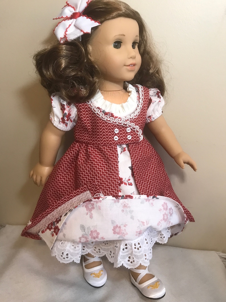 May include: A doll wearing a red and white dress with a floral print. The dress has a white lace trim and a white ruffle collar. The doll is wearing white shoes with gold accents.