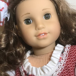 May include: A doll with long brown curly hair, wearing a red and white dress with a white lace trim. The doll has green eyes and a light brown complexion.