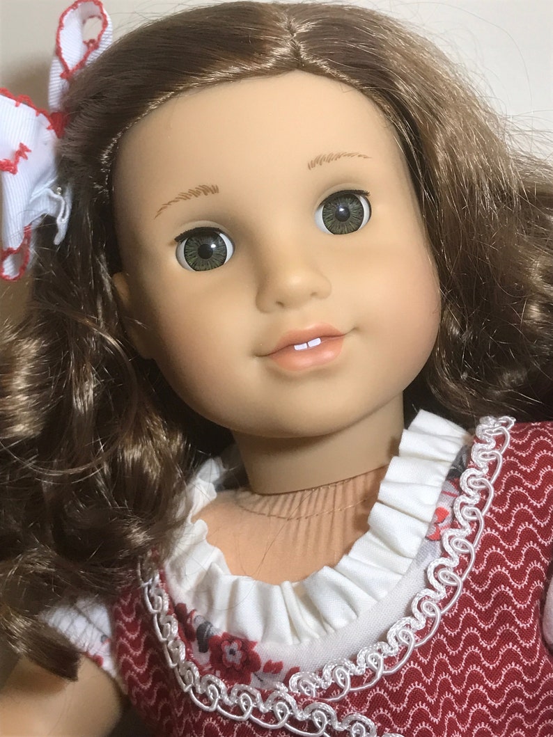 May include: A doll with long brown curly hair, wearing a red and white dress with a white lace trim. The doll has green eyes and a light brown complexion.