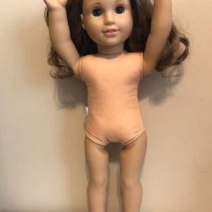 May include: A brown-haired doll with green eyes wearing a light brown sleeveless leotard. The doll is standing with her arms raised above her head.