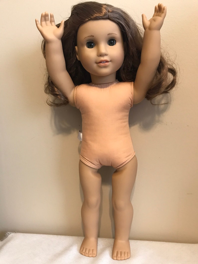May include: A brown-haired doll with green eyes wearing a light brown sleeveless leotard. The doll is standing with her arms raised above her head.