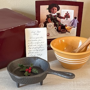 May include: An assortment of kitchen items including a small cast iron pan with a handle and holly berries, a yellow and white striped bowl with a wooden spoon, and a recipe card. A doll and box are in the background.