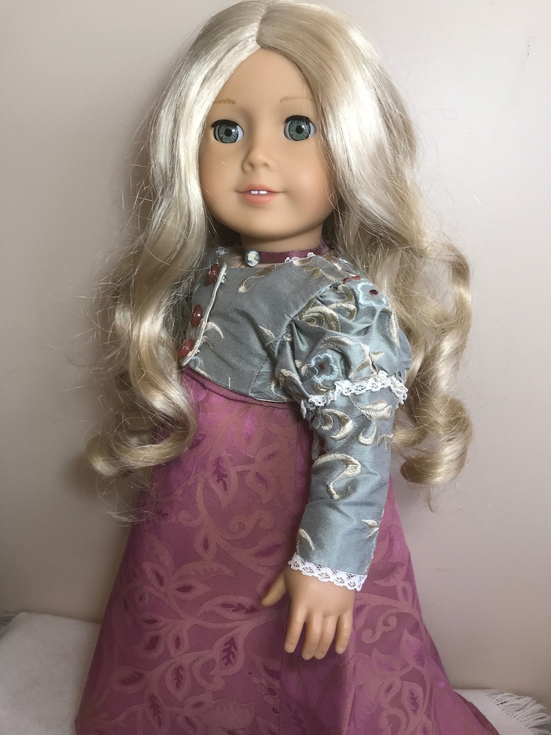 May include: A doll with long blonde hair wearing a green and gold dress with a pink skirt. The doll has green eyes and is smiling.
