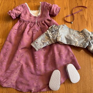 May include: A pink and gold doll dress with a matching jacket and a pair of white shoes. The dress has a lace trim and a floral pattern. The jacket has a gold trim and a floral pattern. The shoes are simple and white.
