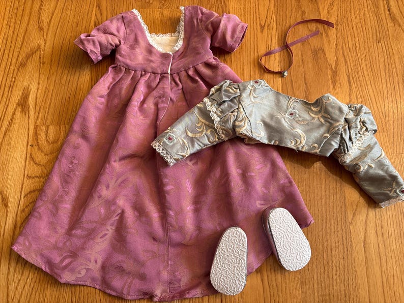 May include: A pink and gold doll dress with a matching jacket and a pair of white shoes. The dress has a lace trim and a floral pattern. The jacket has a gold trim and a floral pattern. The shoes are simple and white.