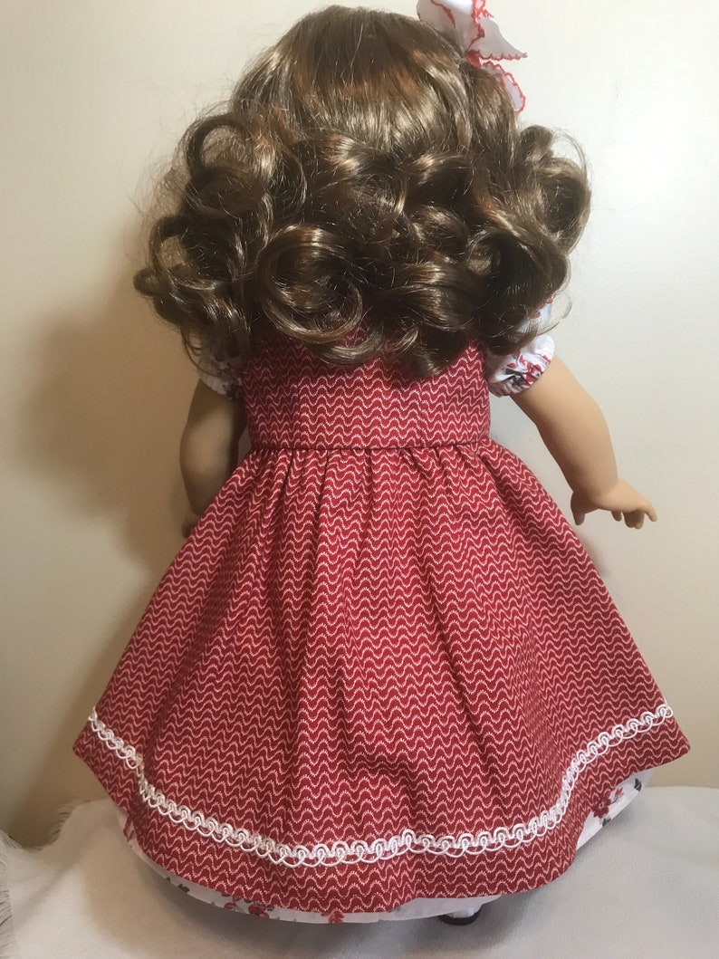 May include: A red dress with white trim and a white and red patterned underskirt. The dress has a scalloped hem and is made of a soft, cotton fabric. The dress is perfect for a doll.